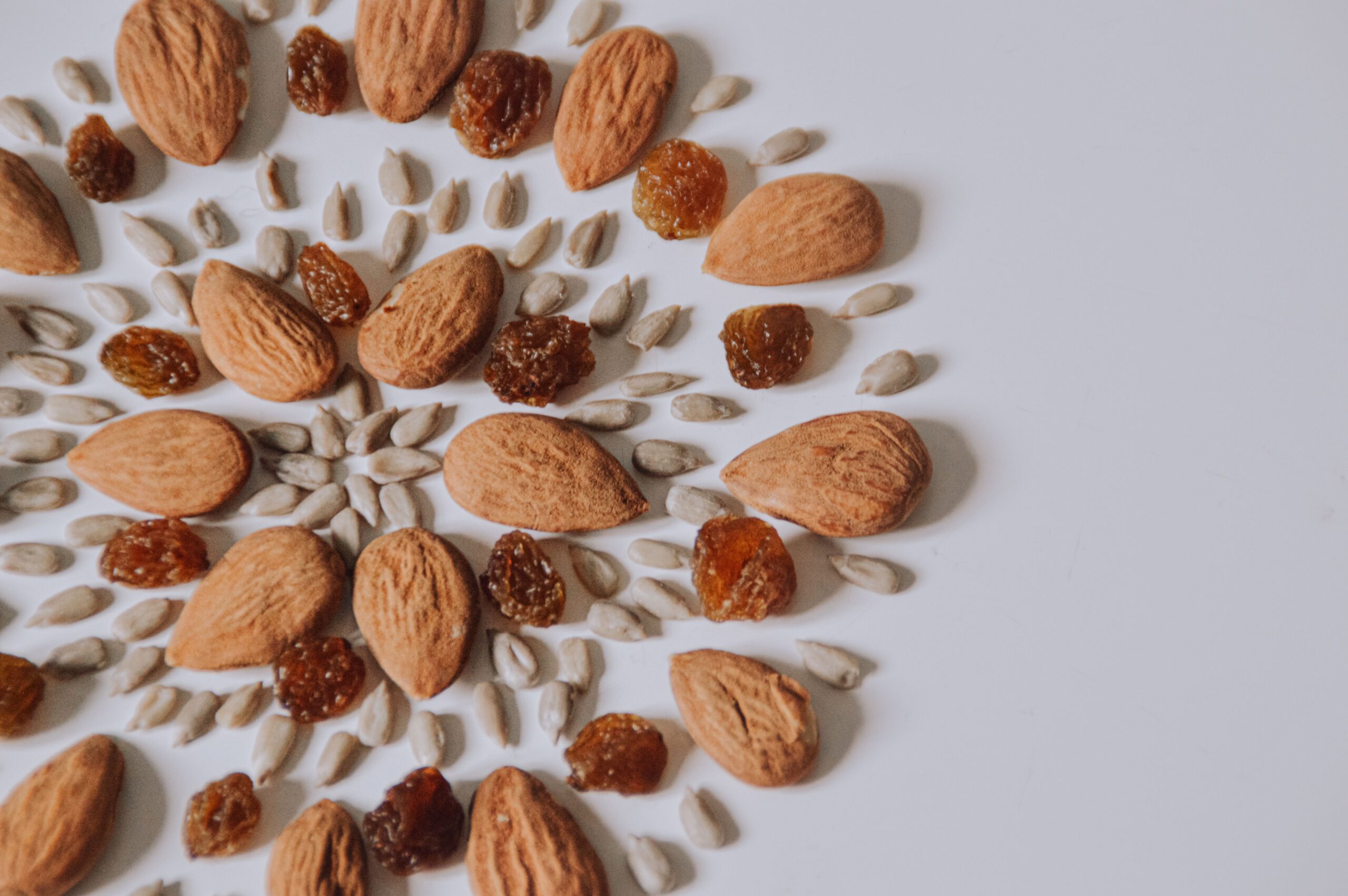 Artistic pattern of almonds, raisins, and seeds on white surface.